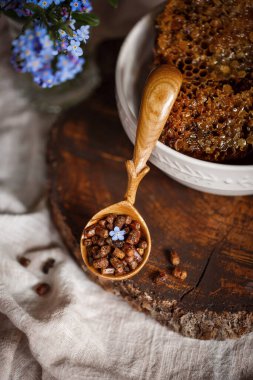 Ahşap kahverengi kaşık arı ürünleri ile Perga arı parşömeninin yanında bal ve ahşap bir kahverengi kesikle keten bir masa örtüsü üzerinde mavi çiçekler ve parşömenin üzerinde bir çiçek.