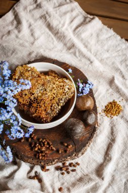 Propolis, parşömen, arı ürünleri, polenlerin yanındaki kahverengi bir ahşap tabakta taraktaki işlenmemiş bal. Masanın üzerinde keten masa örtüsü, mavi çiçeklerin yanında şık bir resim.