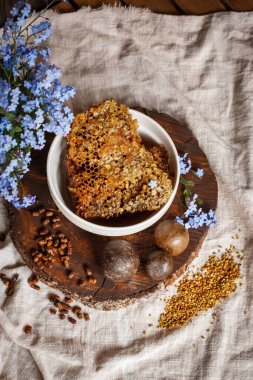Propolis, parşömen, arı ürünleri, polenlerin yanındaki kahverengi bir ahşap tabakta taraktaki işlenmemiş bal. Masanın üzerinde keten masa örtüsü, mavi çiçeklerin yanında şık bir resim.