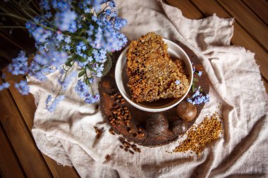 Propolis, parşömen, arı ürünleri, polenlerin yanındaki kahverengi bir ahşap tabakta taraktaki işlenmemiş bal. Masanın üzerinde keten masa örtüsü, mavi çiçeklerin yanında şık bir resim.