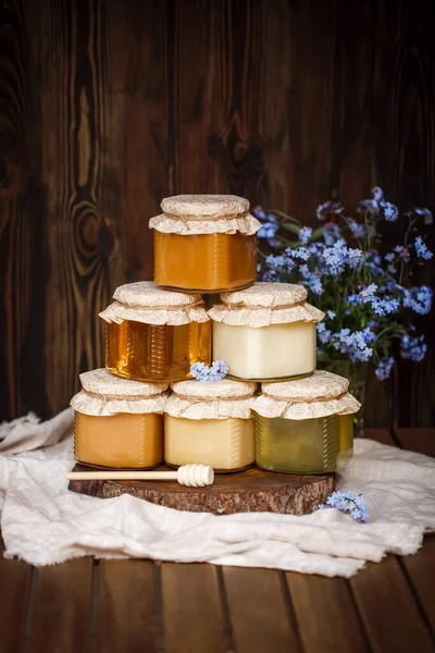 Çiğ organik ballı kavanoz piramidi. Bal sarısı, kahverengi, birbirinin üstünde duruyor. Tahta bir testere üzerinde keten bir masa örtüsü üzerinde kahverengi ahşap bir masada mavi çiçeklerin yanında.