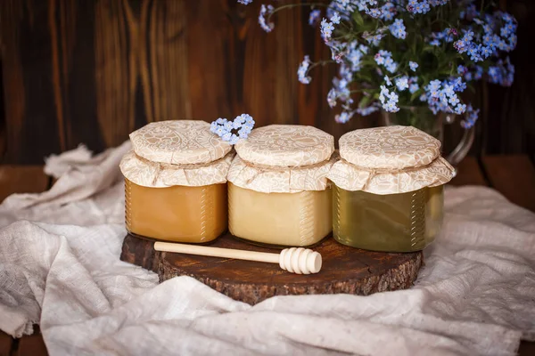Üç kavanoz taze bal farklı renklerde beyaz kahverengi ve sarı bir tahta testerenin üzerinde kahverengi bir masada kahverengi bir fonda mavi çiçeklerin yanında kahverengi bir masa örtüsü.