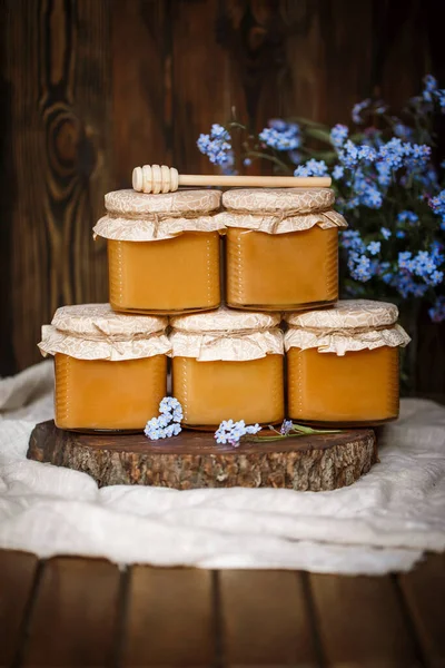 Çiğ organik ballı kavanoz piramidi. Bal sarısı, kahverengi, birbirinin üstünde duruyor. Tahta bir testere üzerinde keten bir masa örtüsü üzerinde kahverengi ahşap bir masada mavi çiçeklerin yanında.