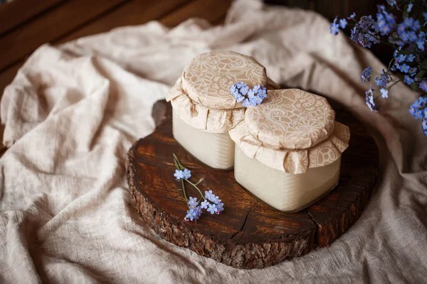 Kavanozlara bağlanmış taze beyaz bal ve kahverengi bir ağacın üzerinde kahverengi bir masa örtüsünün üzerinde güzel bir kapak kahverengi bir masa örtüsü. kalp şeklinde çiçekler.