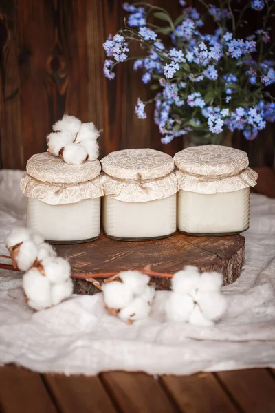 Tahta kahverengi testereyle yan yana duran üç kavanoz beyaz pamuk balı ahşap bir masa örtüsü üzerinde ahşap kahverengi bir masa örtüsü ile pamuk çiçeklerinin yanındaki ahşap kahverengi arka plan üzerinde bir parça ahşap kesilir. 