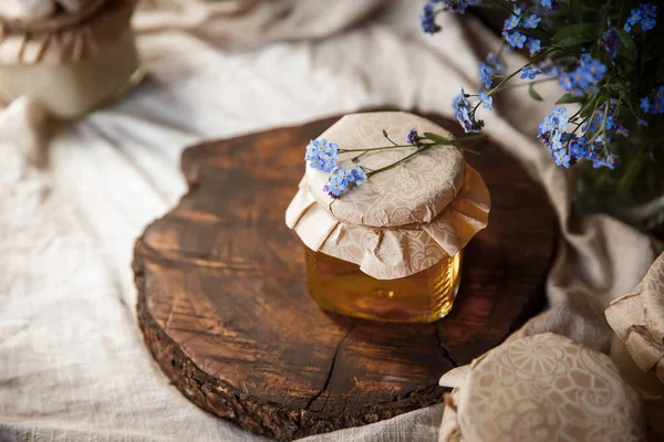  Kahverengi ahşap bir kesikte taze taze sunny bal kavanozu. Kapağının üstünde çiçekler vardı. Kapağı iple sarılmış. Kahverengi bir sehpanın üzerindeki keten kumaş üzerinde kahverengi bir ahşap arka planda mavi çiçeklerin yanında. Güzel, şık, lezzetli, tatlı, lezzetli kırsal... 