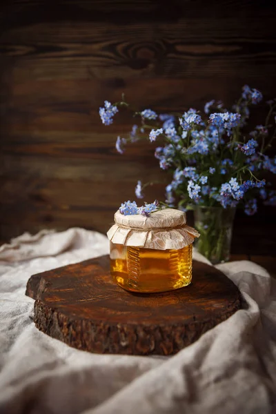 Kahverengi ahşap bir kesikte taze sunny ham bal kavanozu. Kapağının üstünde çiçekler vardı. Kapağı iple sarılmış. Kahverengi bir sehpanın üzerindeki keten kumaş üzerinde kahverengi bir ahşap arka planda mavi çiçeklerin yanında. Güzel, şık, lezzetli, tatlı, lezzetli kırsal... 
