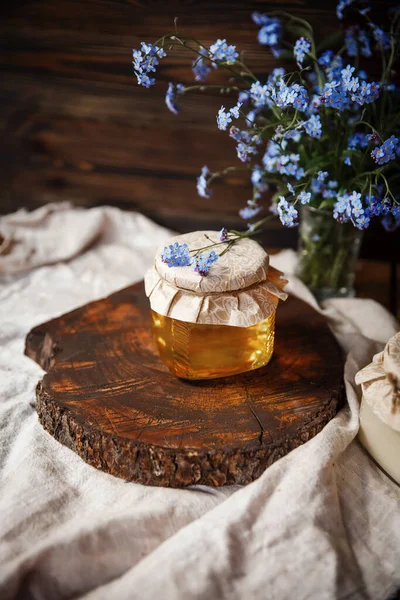  Kahverengi ahşap bir kesikte taze taze sunny bal kavanozu. Kapağının üstünde çiçekler vardı. Kapağı iple sarılmış. Kahverengi bir sehpanın üzerindeki keten kumaş üzerinde kahverengi bir ahşap arka planda mavi çiçeklerin yanında. Güzel, şık, lezzetli, tatlı, lezzetli kırsal... 