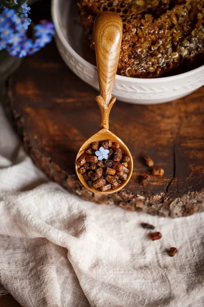 Ahşap kahverengi kaşık arı ürünleri ile Perga arı parşömeninin yanında bal ve ahşap bir kahverengi kesikle keten bir masa örtüsü üzerinde mavi çiçekler ve parşömenin üzerinde bir çiçek.
