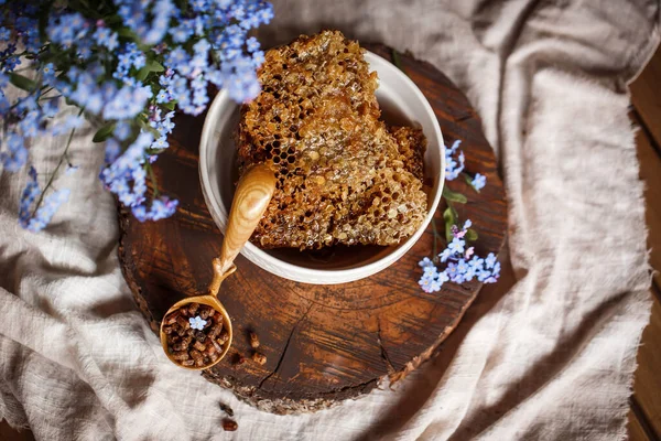 Ahşap kahverengi kaşık arı ürünleri ile Perga arı parşömeninin yanında bal ve ahşap bir kahverengi kesikle keten bir masa örtüsü üzerinde mavi çiçekler ve parşömenin üzerinde bir çiçek.