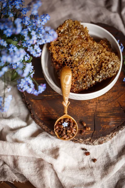 Ahşap kahverengi kaşık arı ürünleri ile Perga arı parşömeninin yanında bal ve ahşap bir kahverengi kesikle keten bir masa örtüsü üzerinde mavi çiçekler ve parşömenin üzerinde bir çiçek.