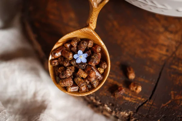 Ahşap kahverengi kaşık arı ürünleri ile Perga arı parşömeninin yanında bal ve ahşap bir kahverengi kesikle keten bir masa örtüsü üzerinde mavi çiçekler ve parşömen üzerinde bir çiçek.