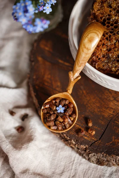Ahşap kahverengi kaşık arı ürünleri ile Perga arı parşömeninin yanında bal ve ahşap bir kahverengi kesikle keten bir masa örtüsü üzerinde mavi çiçekler ve parşömenin üzerinde bir çiçek.