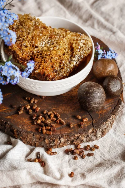 Propolis, parşömen, arı ürünleri, polenlerin yanındaki kahverengi bir ahşap tabakta taraktaki işlenmemiş bal. Masanın üzerinde keten masa örtüsü, mavi çiçeklerin yanında şık bir resim.