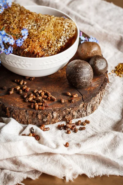 Propolis, parşömen, arı ürünleri, polenlerin yanındaki kahverengi bir ahşap tabakta taraktaki işlenmemiş bal. Masanın üzerinde keten masa örtüsü, mavi çiçeklerin yanında şık bir resim.