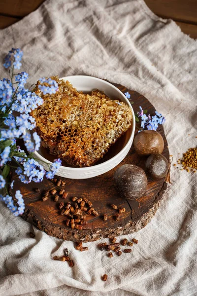 Propolis, parşömen, arı ürünleri, polenlerin yanındaki kahverengi bir ahşap tabakta taraktaki işlenmemiş bal. Masanın üzerinde keten masa örtüsü, mavi çiçeklerin yanında şık bir resim.