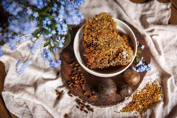 Propolis, parşömen, arı ürünleri, polenlerin yanındaki kahverengi bir ahşap tabakta taraktaki işlenmemiş bal. Masanın üzerinde keten masa örtüsü, mavi çiçeklerin yanında şık bir resim.