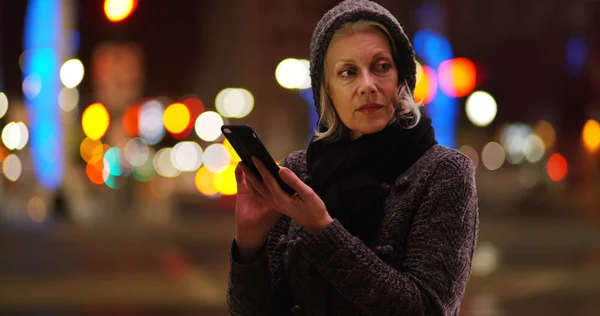 White old woman in sweater and scarf using phone in urban setting at night