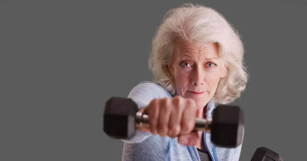 Determined senior woman exercising with dumbbells on solid gray backdrop