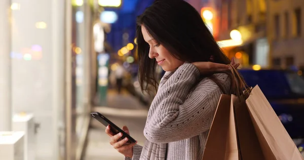 Beautiful female carrying shopping bags and texting on city street at night