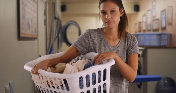 Serious young woman holding basket of dirty clothes in laundry room