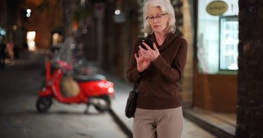 Mature woman tourist checking map or rideshare app on cell phone at night clipart