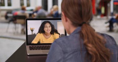 Millennial brunette video chatting with Hispanic friend on her computer at cafe clipart