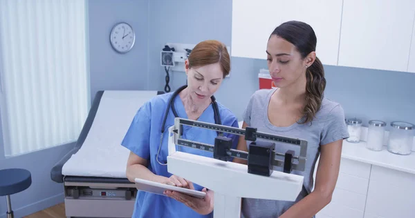 Charming middle aged nurse using scale to measure weight of young latina patient
