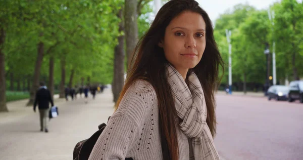 Millennial woman in striped scarf smiling at camera near public park in London