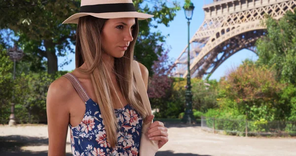 Pretty lady in floral romper wearing fedora and holding bag near Eiffel Tower