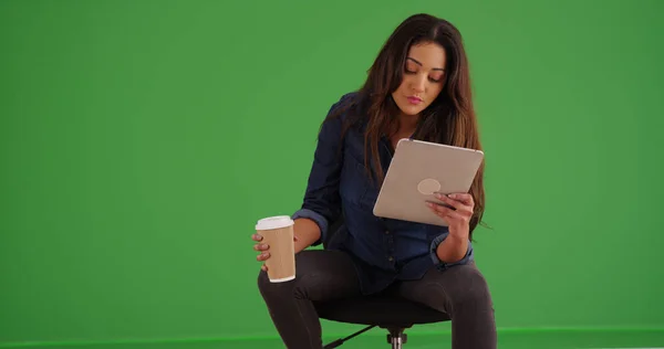 Latina with coffee using tablet sitting in swivel chair on green screen