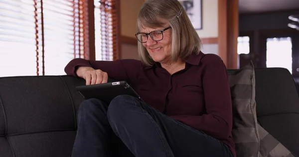 在客厅里使用手持平板电脑坐在沙发上的退休成熟妇女 — 图库照片