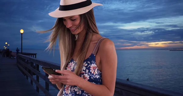 Happy woman in floral romper and fedora texting on phone on pier in the evening