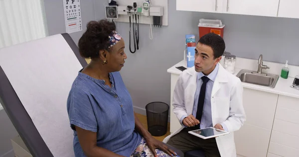 向办公室里的年轻男性西班牙裔医生解释健康问题的老黑人妇女 — 图库照片