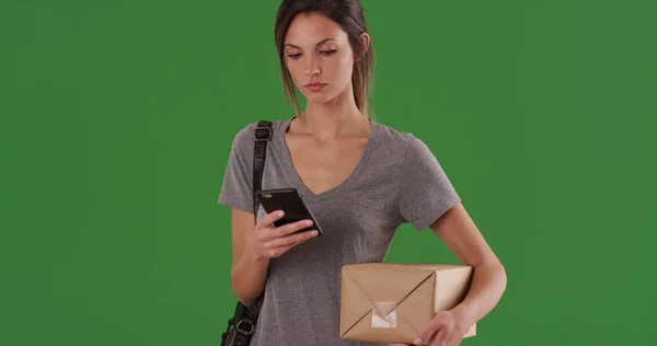 Woman checks shipping service app on phone while holding package on greenscreen