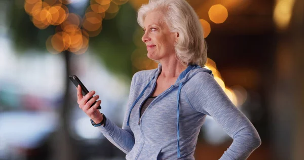 Happy old woman in hoodie holding mobile device and looking off-screen outdoors