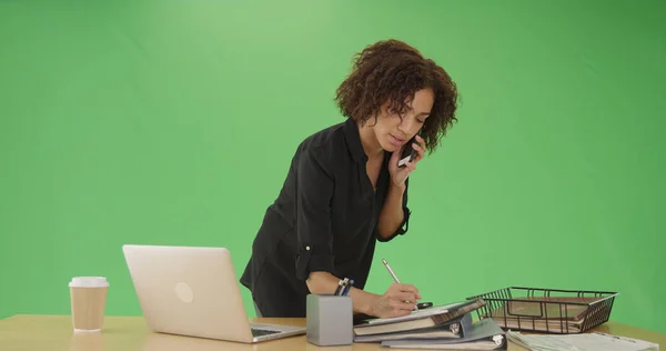 Young black office worker writing notes and using phone on green screen