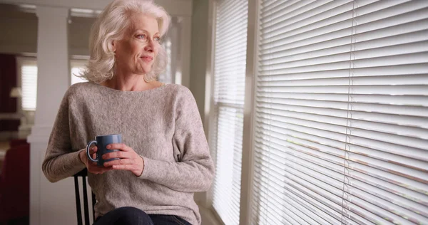 带着咖啡杯的老年白人女士坐在家里 望着窗外 — 图库照片