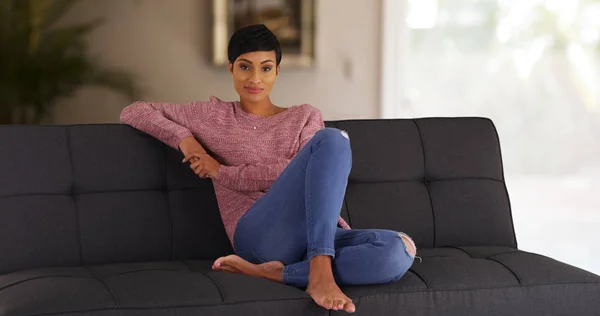 セーターとジーンズのカメラで笑顔のソファーでのんびりで黒人女性の肖像画 — ストック写真