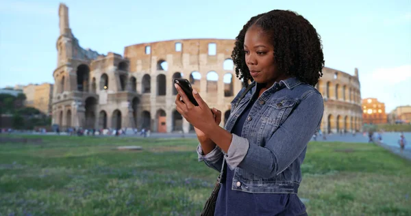 Black woman on vacation in Rome reading text message from friend by Colisseum