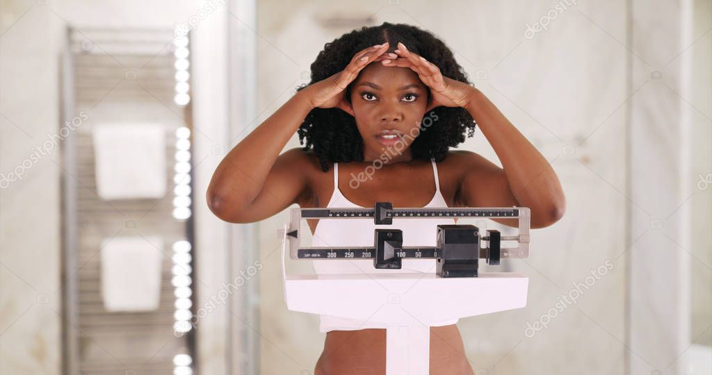 Young black woman weighs herself on scale frowning and shaking her head