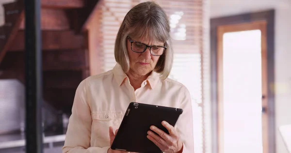 Independent senior woman using tablet device to browse online inside modern home