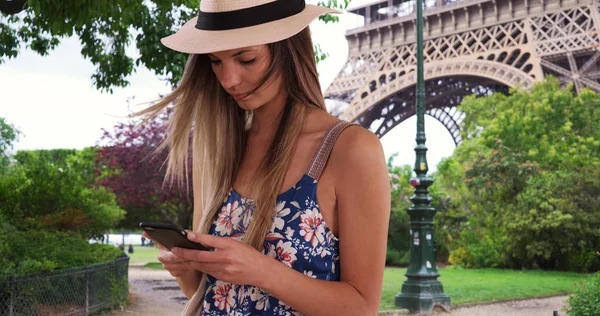 Millennial girl in romper and fedora texting on phone by Eiffel Tower in Paris