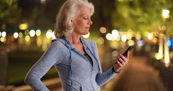 Elder female in blue hoodie sending text on smartphone outside at night
