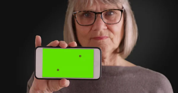 灰色の背景に緑色の画面で携帯電話を保持している高齢者の白人女性 — ストック写真