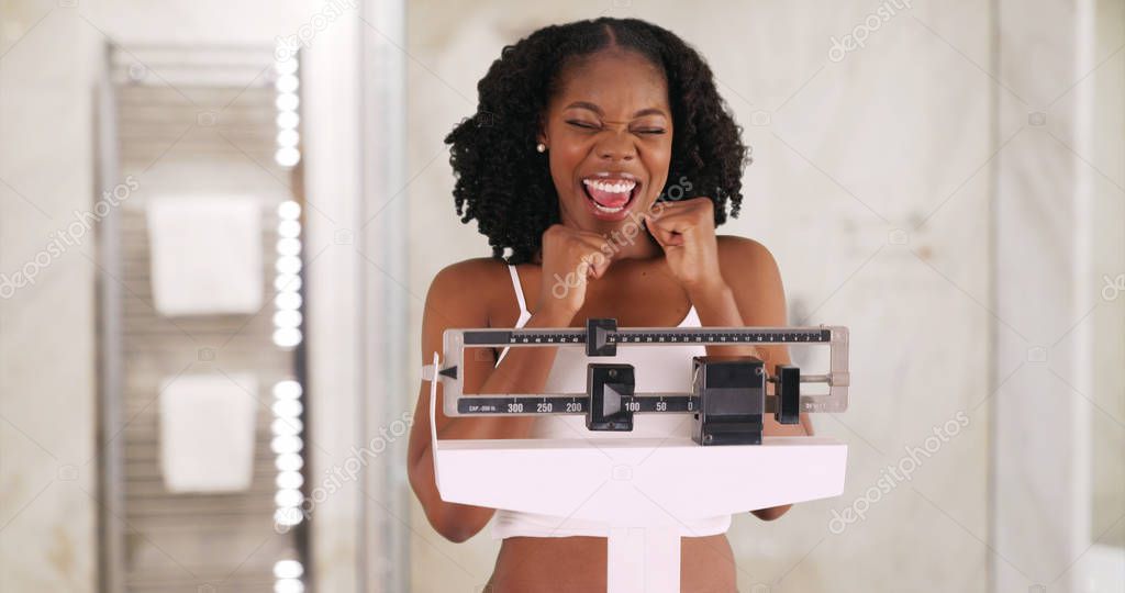 Beautiful black woman happily weighs herself on scale in bathroom