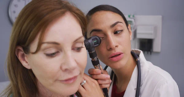 Female patient having her ear examined with otoscope by young latina doctor