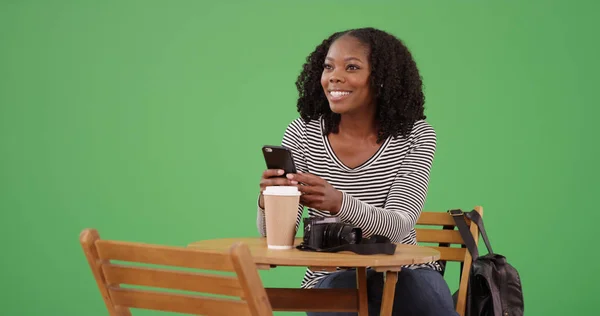 Smiling black female using mobile device while sitting at cafe on green screen