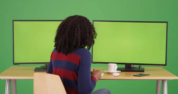 Black woman sitting by computers with green screens on display on green screen.