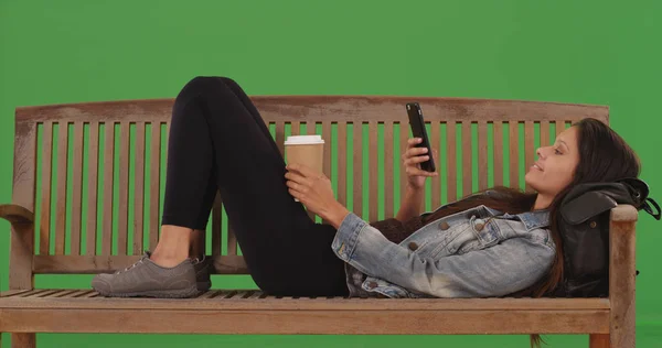 Pretty young woman lying down on bench using smartphone on green screen